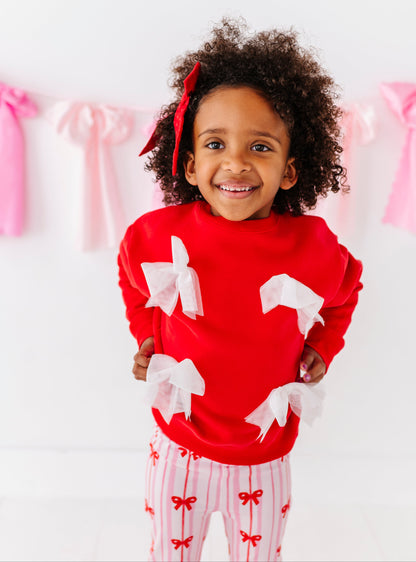 Child wearing a red crewneck sweater with white bows and flare bamboo pants with red bows, smiling.