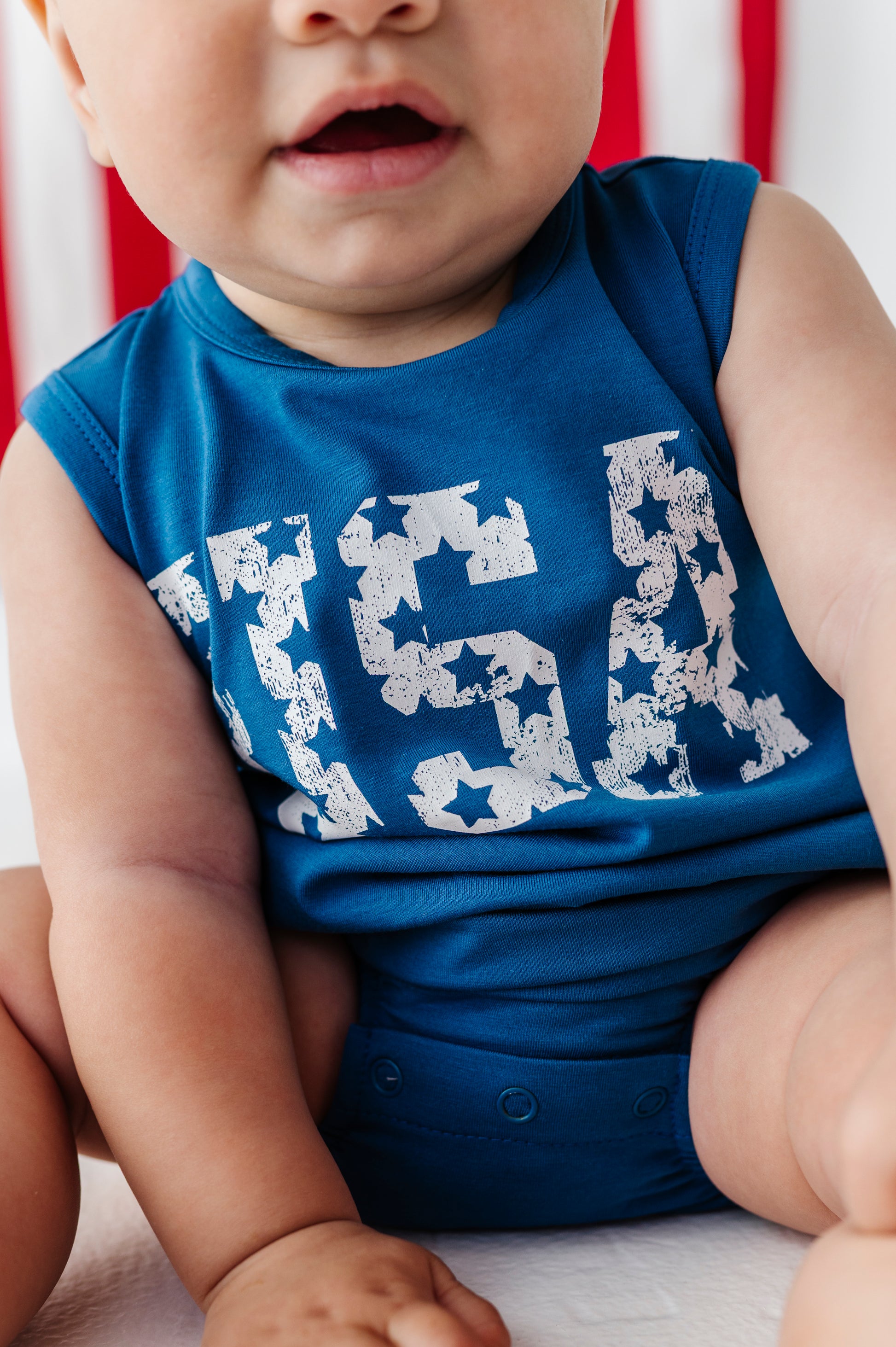 A close-up of a baby wearing a sleeveless royal blue bodysuit with a distressed white 'USA' graphic across the chest, sitting against a soft red and white striped background.