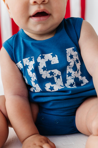 A close-up of a baby wearing a sleeveless royal blue bodysuit with a distressed white 'USA' graphic across the chest, sitting against a soft red and white striped background.