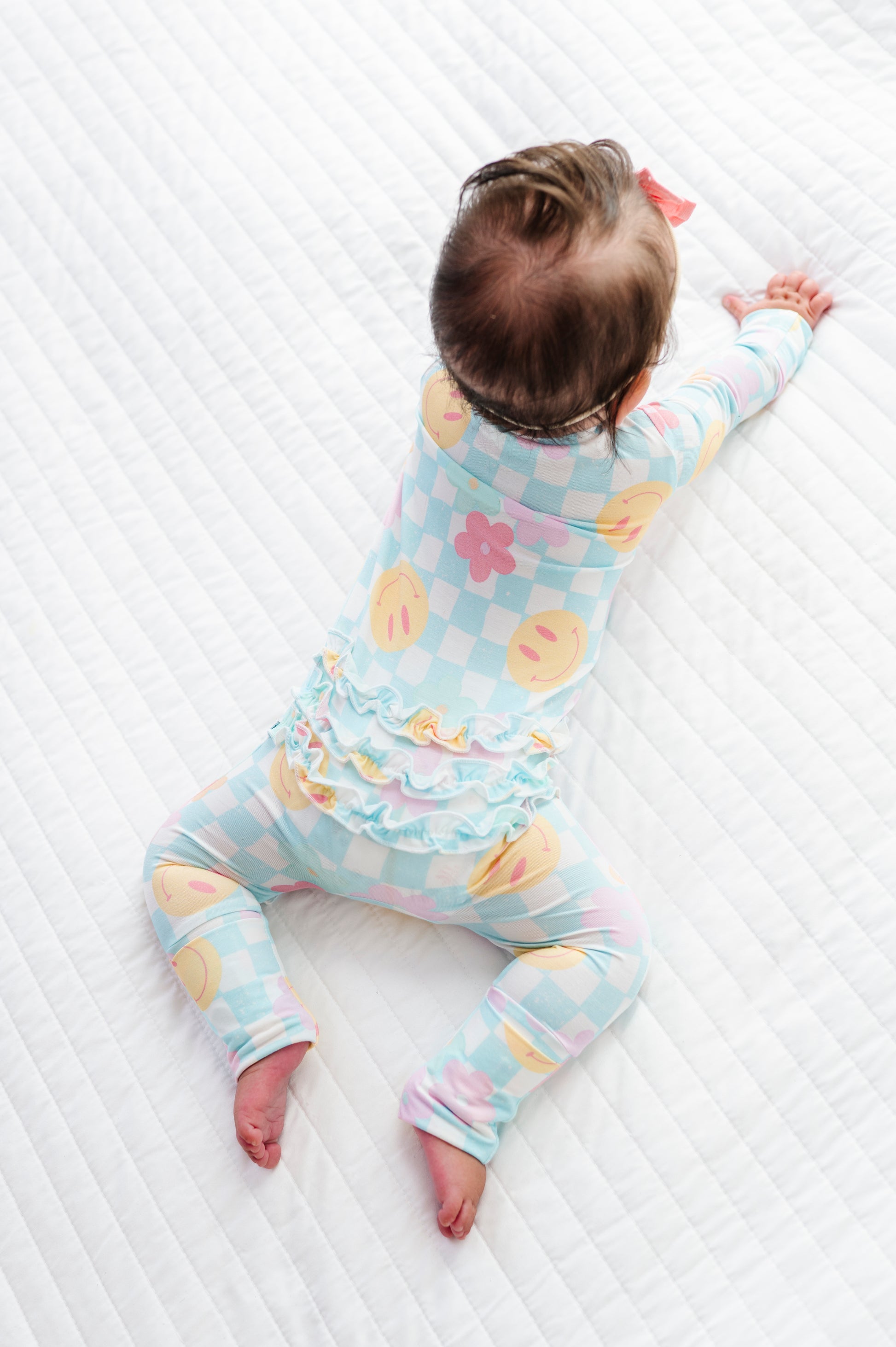 A baby lying on a white quilted mattress, wearing a pastel blue, pink, and yellow checkered long-sleeve romper with a ruffled waistband.