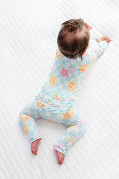 A baby lying on a white quilted mattress, wearing a pastel blue, pink, and yellow checkered long-sleeve romper with a ruffled waistband.