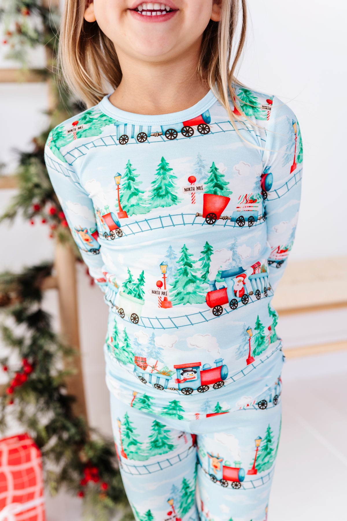 A child wearing a light blue long-sleeve bamboo pajama set with a Christmas train and tree print, standing indoors near a decorated Christmas tree.