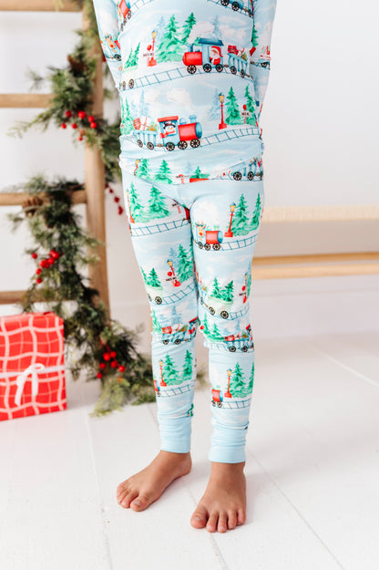 A child wearing light blue Christmas-themed bamboo pajama pants with a red train and green pine tree pattern, standing barefoot in a decorated room.