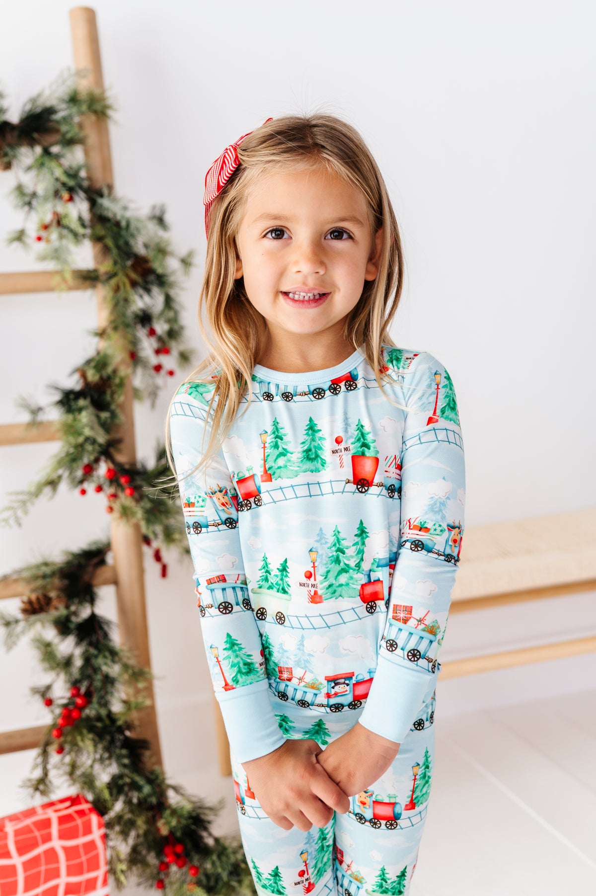 A young girl with blonde hair wearing a red bow headband and a light blue long-sleeve bamboo pajama set with a Christmas train and tree pattern, standing in front of a decorated wooden ladder and a white wall.