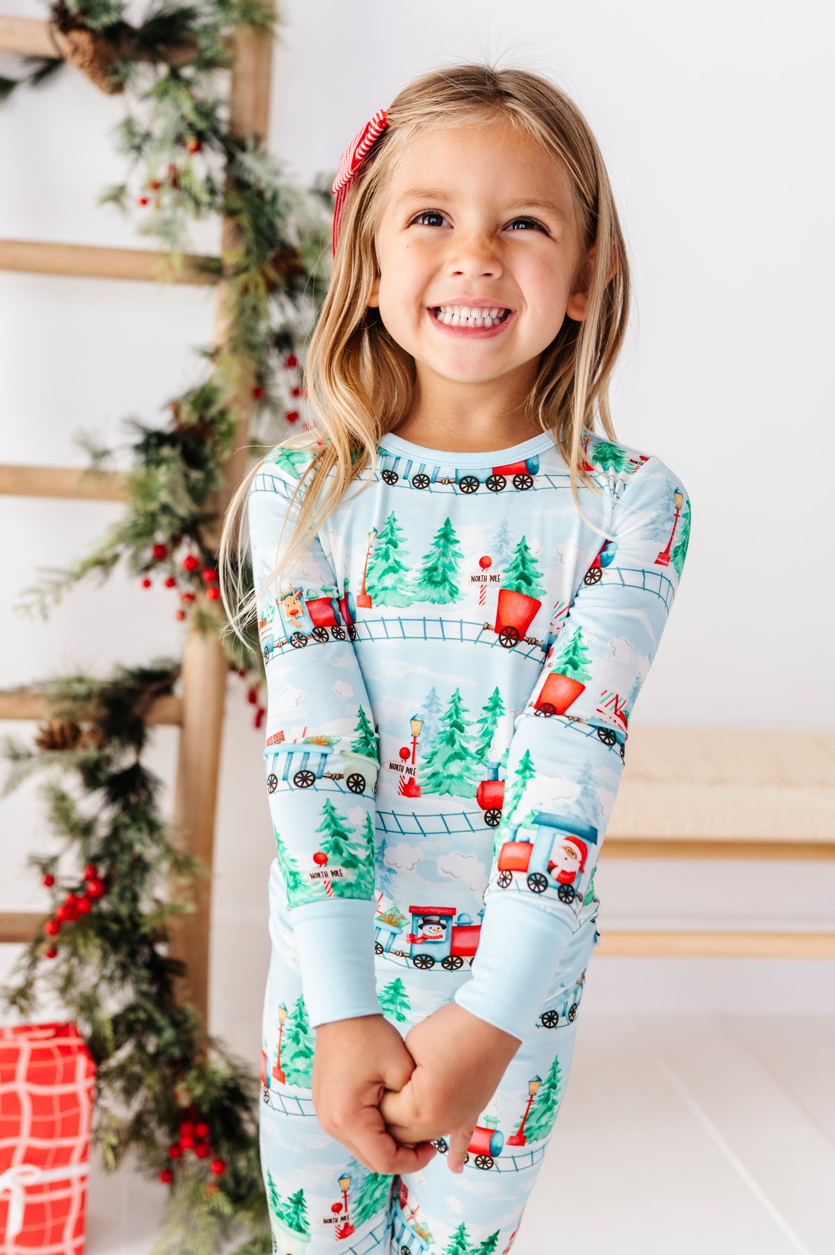 A young girl smiling while wearing a light blue long-sleeve bamboo pajama set with a Christmas train and Santa pattern, posed in front of a decorated holiday tree and wooden ladder.