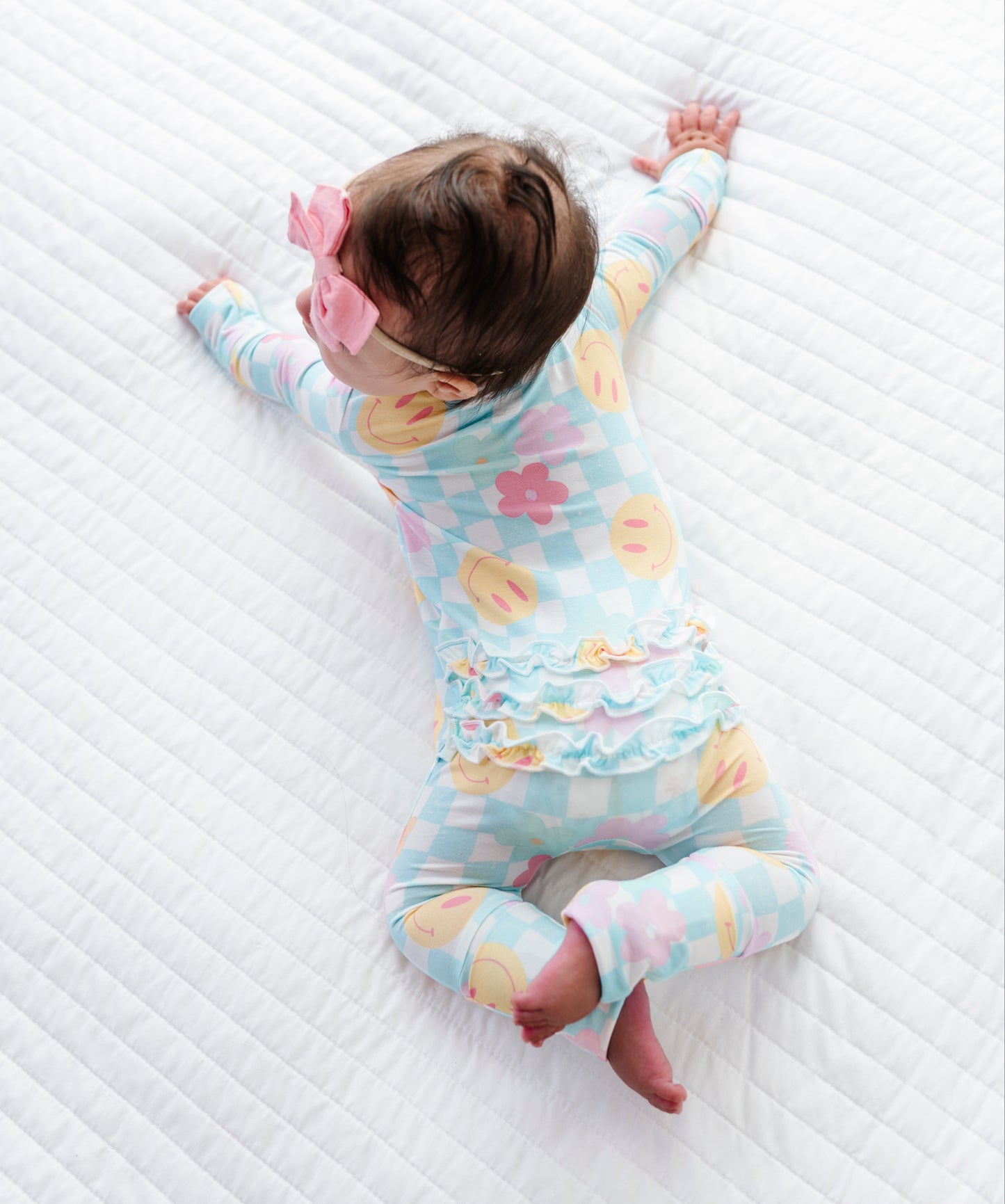 A baby lying on a white quilted mattress, wearing a pastel blue and pink checkered footed sleeper with a ruffled waistband and a pink bow in their hair.