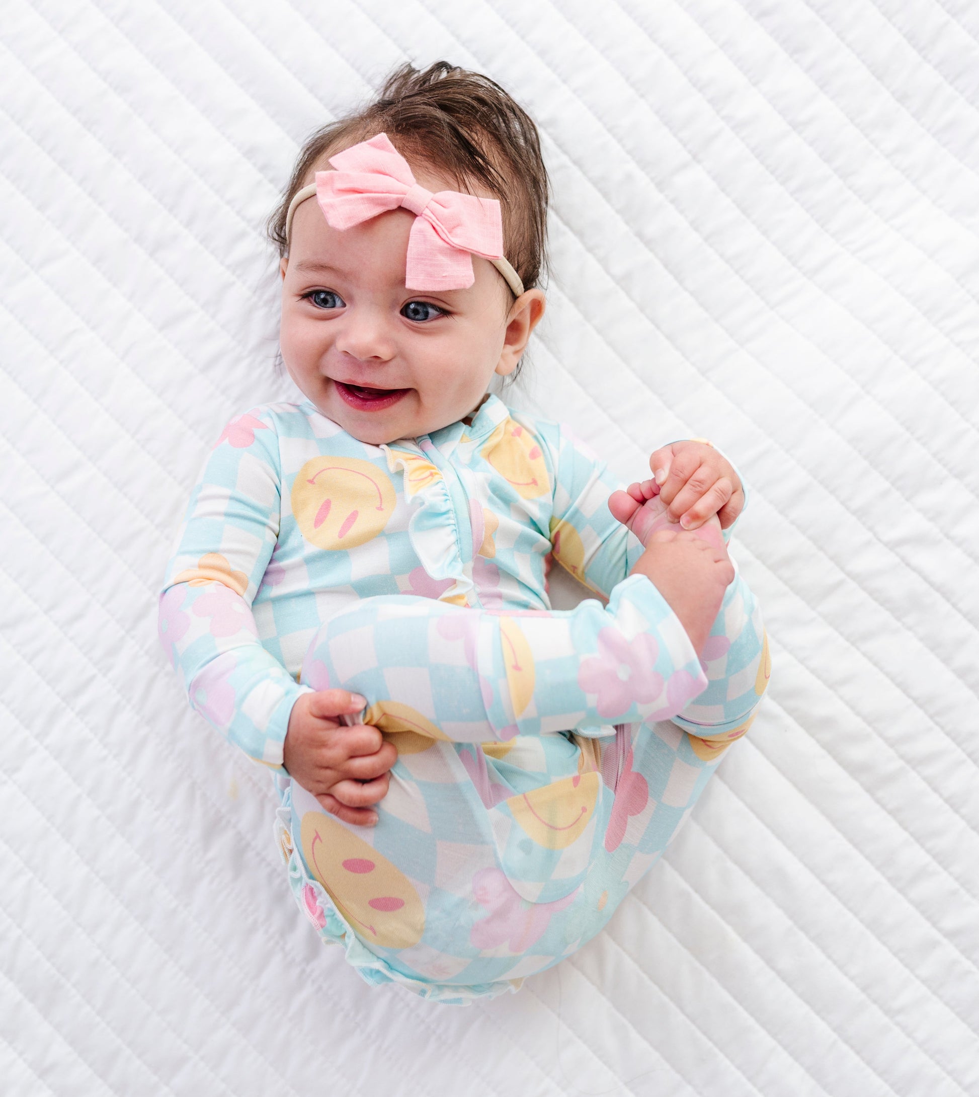 A baby lying on a white quilted blanket, wearing a light blue and pink checkered long-sleeve romper with yellow polka dots, a pink bow headband, and smiling.