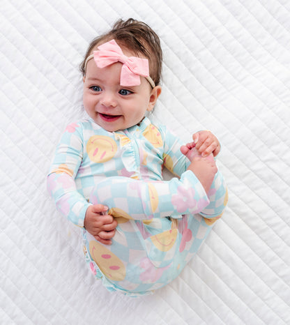 A baby lying on a white quilted blanket, wearing a light blue and pink checkered long-sleeve romper with yellow polka dots, a pink bow headband, and smiling.