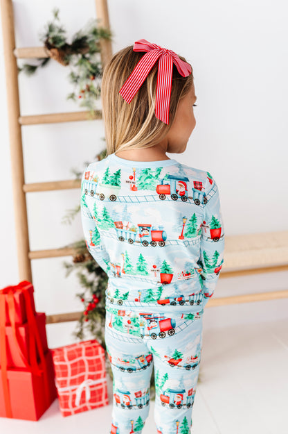 A child wearing a light blue long-sleeve bamboo pajama set with a Christmas train and tree print, shown from the back with a red striped bow in their hair. The background features a wooden ladder, a decorated Christmas tree, and wrapped gifts.