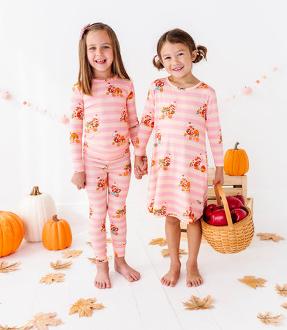 Two young girls standing barefoot in a bright white studio, wearing matching pink long-sleeve pajamas with a fall harvest print. One girl wears a long-sleeve dress version, and the other wears a matching long-sleeve top and pants set.
