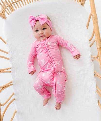 Baby in a pink outfit with a bow lying on a white cushion in a wicker chair.