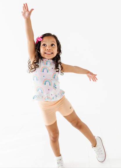 Child wearing a bamboo cotton peplum top with rainbow watercolor patterns and peach bamboo biker shorts on a white background