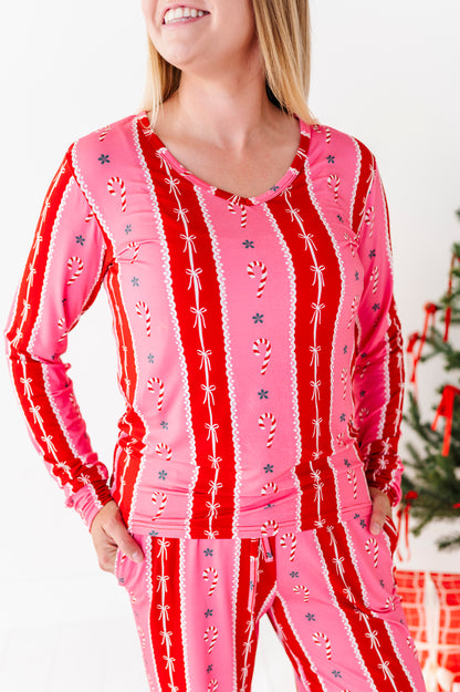 A woman wearing a festive holiday bamboo pajama set with a pink and red striped design featuring candy cane and snowflake patterns, set against a bright white background with a Christmas tree visible on the right.