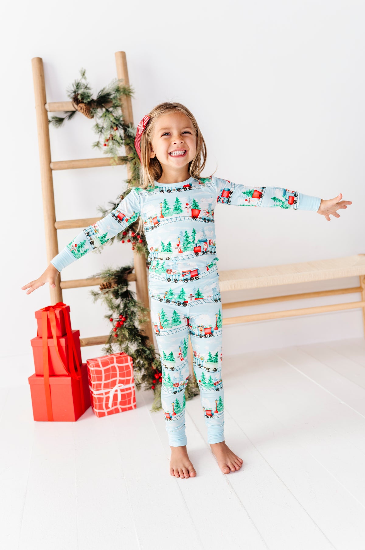 A young girl stands barefoot in a bright room wearing a light blue long-sleeve bamboo pajama set with a Christmas print featuring Santa Claus, trees, and gifts. She is posed with arms outstretched, next to a wooden ladder decorated with greenery and wrapped presents.