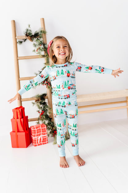 A young girl stands barefoot in a bright room wearing a light blue long-sleeve bamboo pajama set with a Christmas print featuring Santa Claus, trees, and gifts. She is posed with arms outstretched, next to a wooden ladder decorated with greenery and wrapped presents.