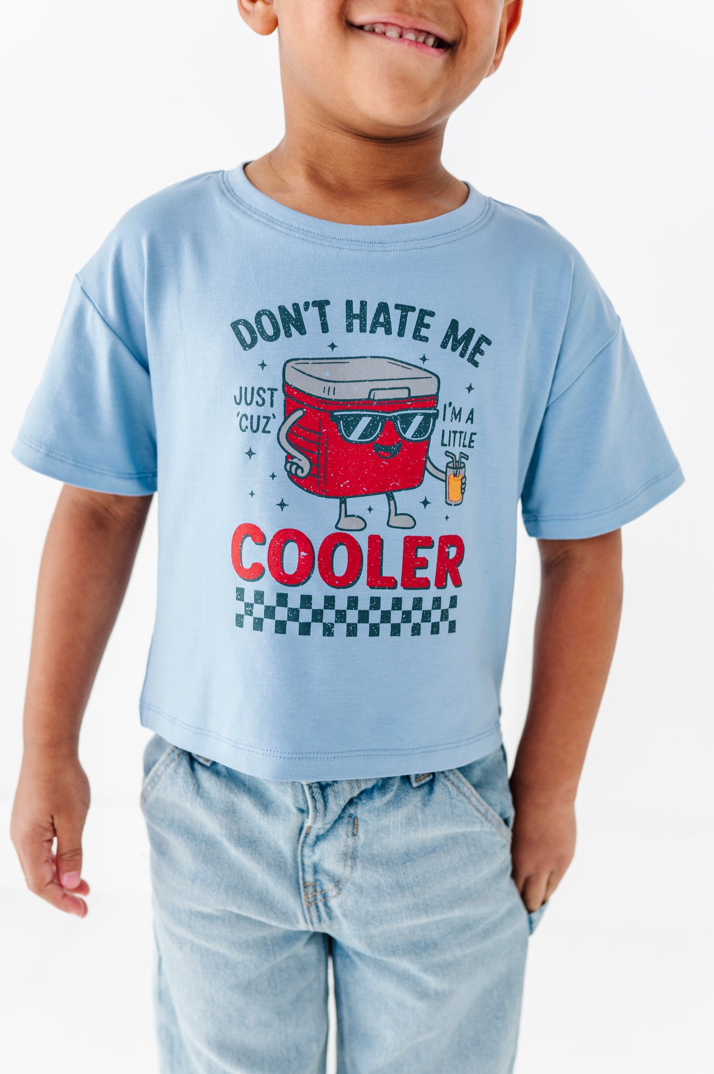 A child wearing a light blue graphic t-shirt with a red cooler character and the text 'DON'T HATE ME COOLER', paired with light-wash jeans against a plain white background.