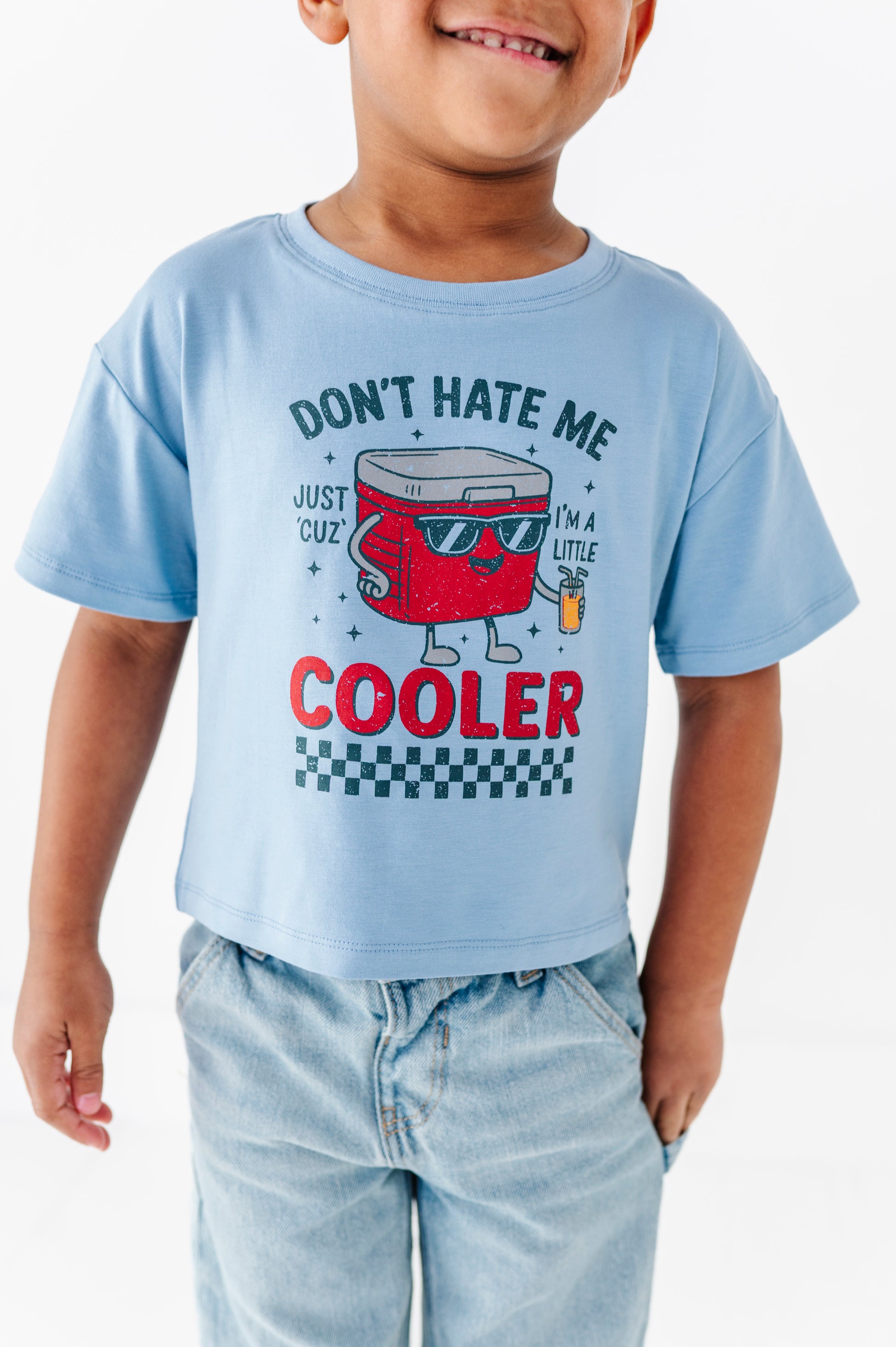 A child wearing a light blue graphic t-shirt with a red cooler character and the text 'DON'T HATE ME COOLER', paired with light-wash jeans against a plain white background.