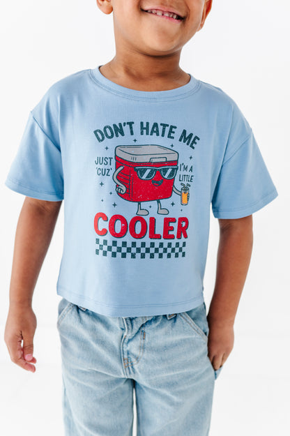 A child wearing a light blue graphic t-shirt with a red cooler character and the text 'DON'T HATE ME COOLER', paired with light-wash jeans against a plain white background.