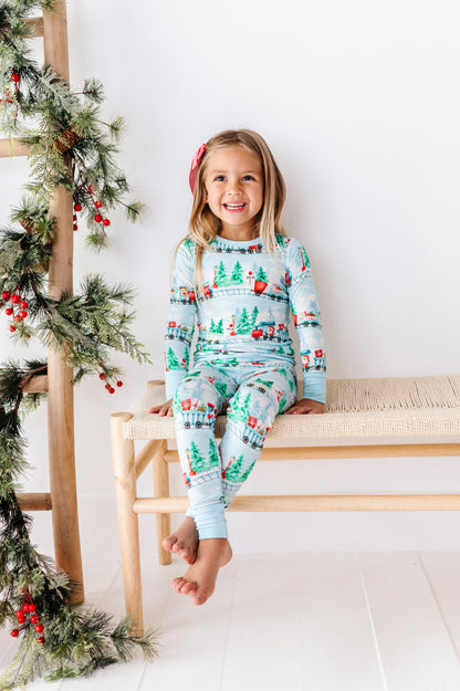 A young girl with blonde hair sits on a light wood bench wearing a light blue long-sleeve bamboo pajama set with a Christmas tree and snowman pattern. The background is a clean white wall with a wooden ladder decorated with green pine garlands and red berries on the left.