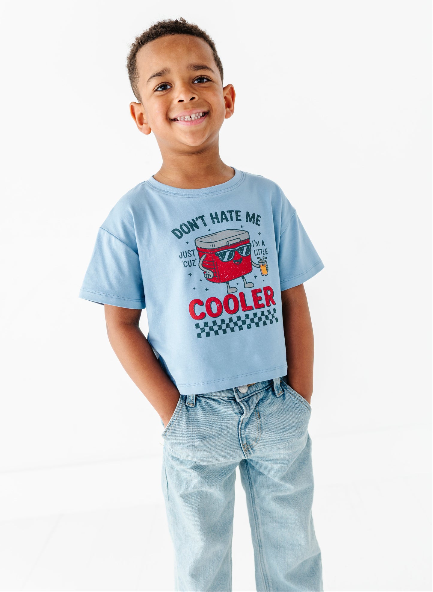 A young boy smiling and wearing a light blue graphic t-shirt with a red cooler design and the text 'DON'T HATE ME JUST TOO MUCH COOLER', paired with light-wash jeans against a white studio background.