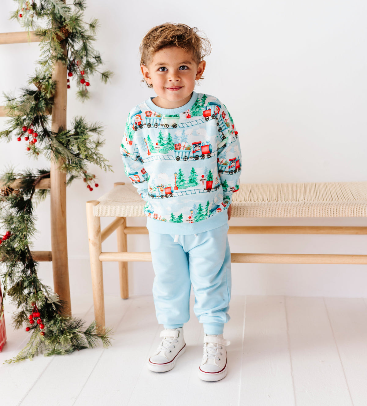 A young child wearing a festive light blue Christmas crewneck sweatshirt with a holiday village print, matching light blue sweatpants, and white sneakers, standing in a bright room with a wooden bench and a ladder decorated with green garlands.