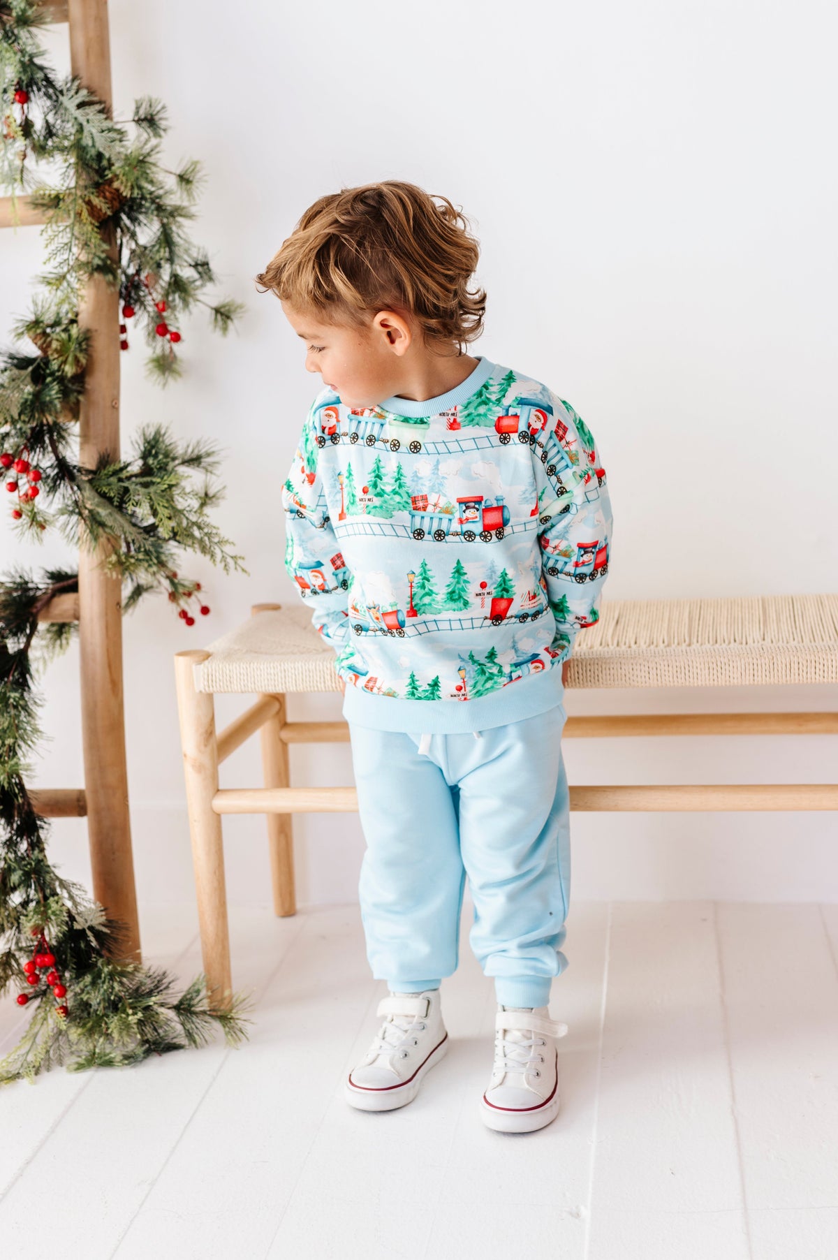 A young child wearing a light blue Christmas-themed crewneck sweatshirt and matching sweatpants, standing in a bright room with a wooden bench and a ladder decorated with green garlands and red berries.