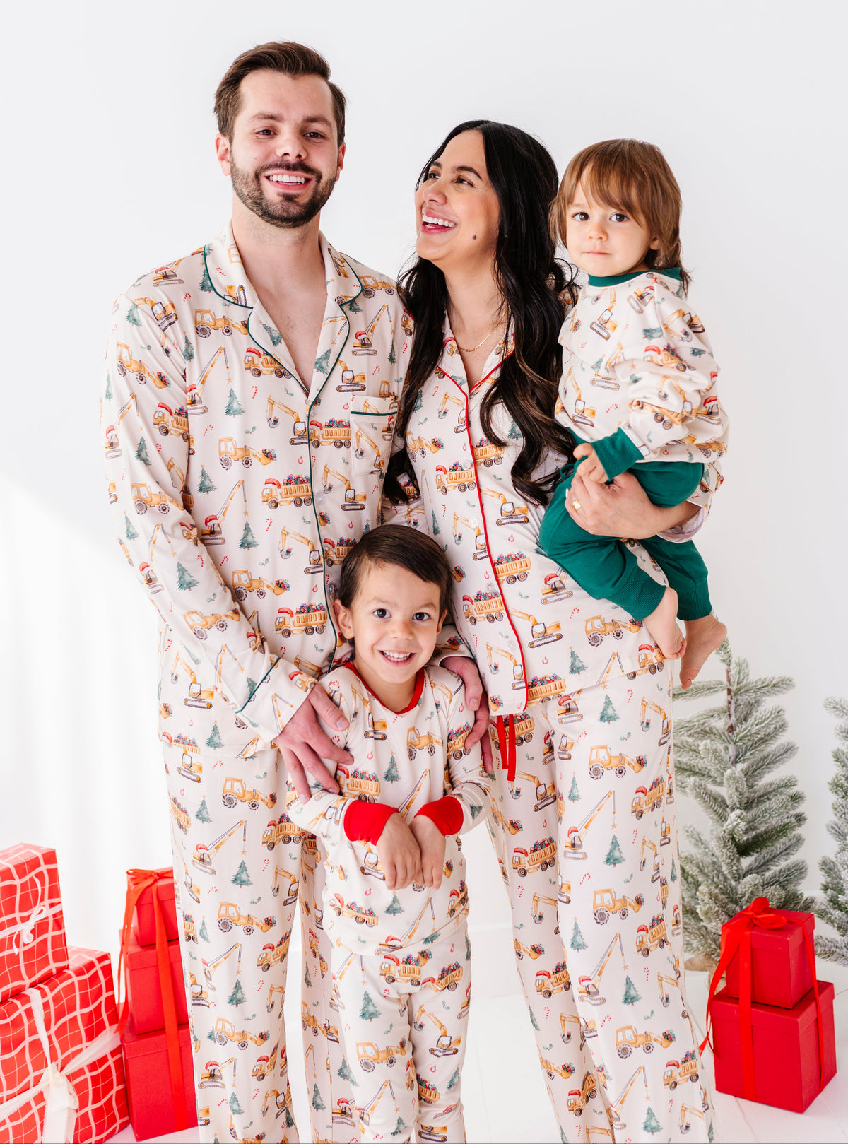 A family wearing long-sleeve bamboo pajama sest with a colorful holiday construction theme print, featuring a cream base with orange, green, and yellow graphics, and dark green piping. 