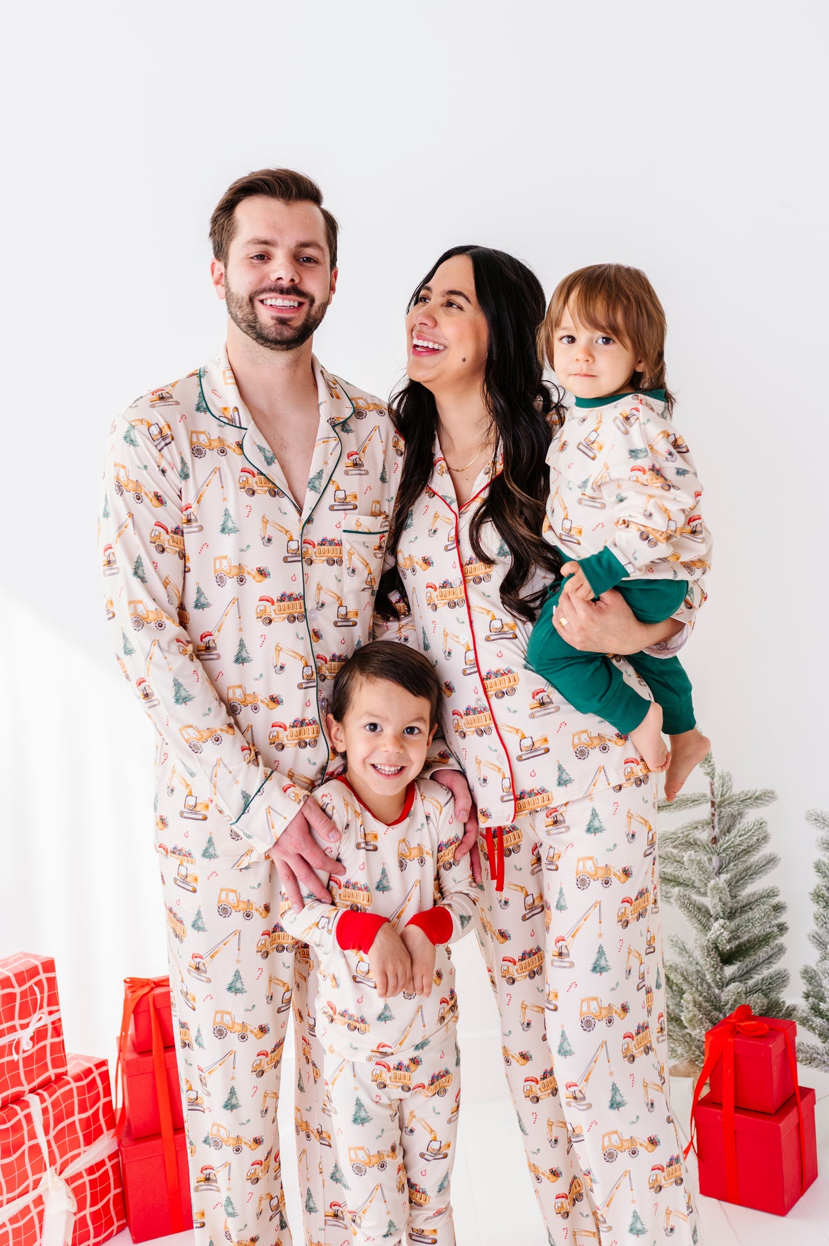 Mom, dad, and two boys wearing matching Christmas construction-themed bamboo pajamas, standing in front of a Christmas tree and a pile of presents 