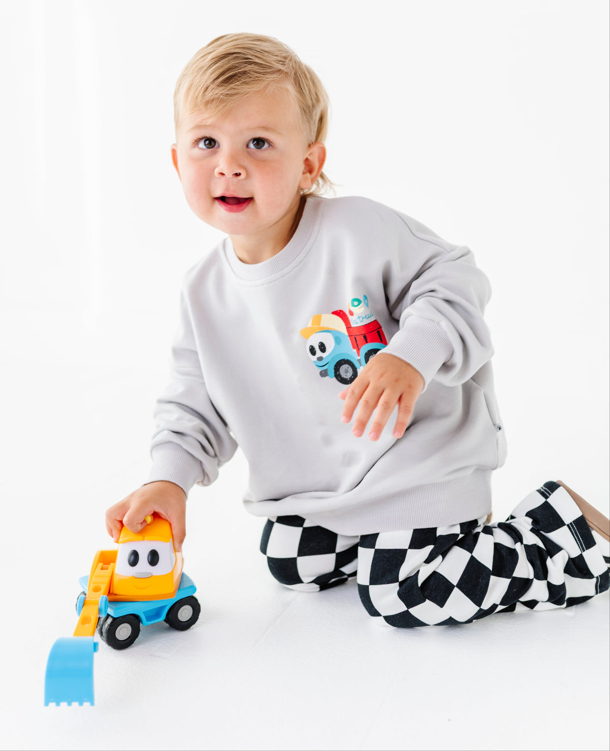 A toddler kneeling on a white studio floor, wearing a light gray sweatshirt with a colorful dump truck graphic and black-and-white checkered pants, while holding a small yellow and blue toy excavator.