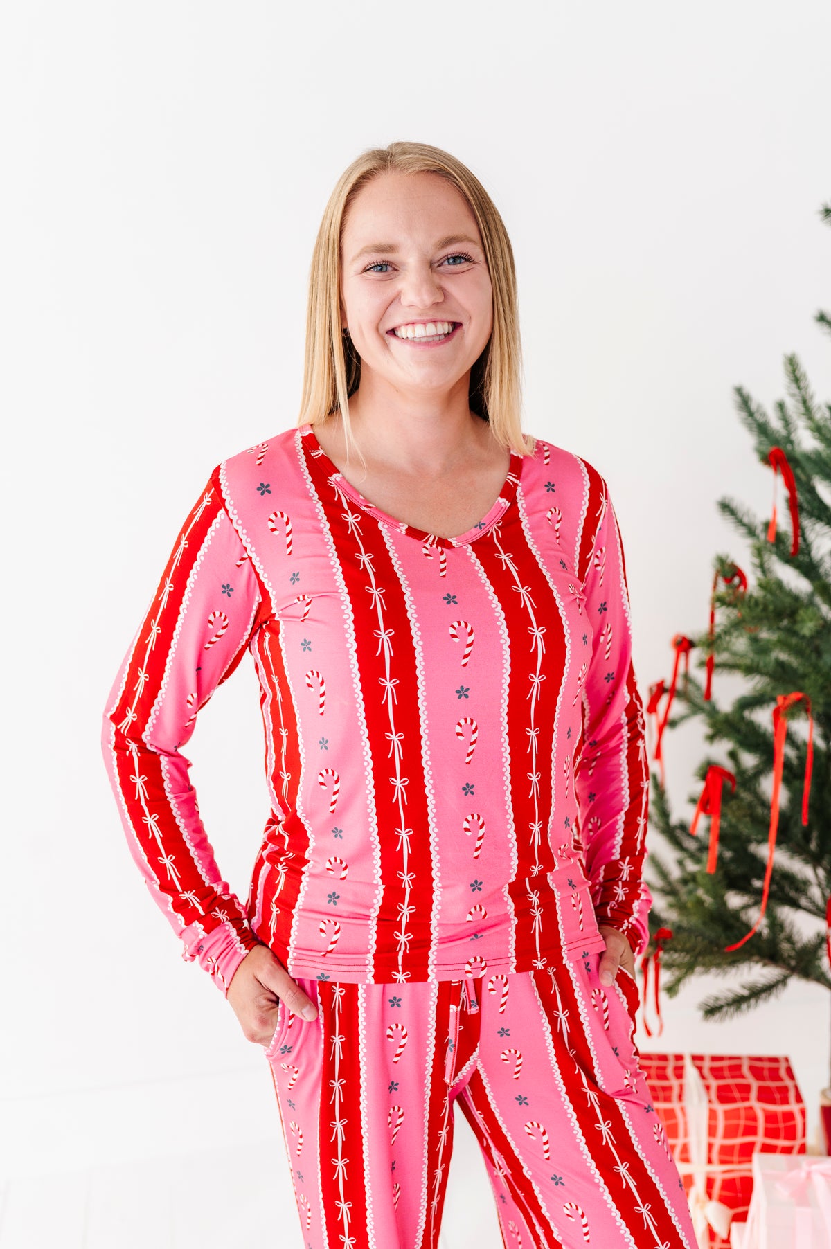 A smiling blonde woman wearing a festive pink and red long-sleeve bamboo pajama set with vertical candy cane and snowflake patterns, standing next to a decorated Christmas tree.