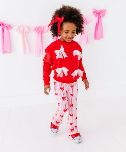 Child wearing a red sweater with white tulle bows and pink stripe bamboo pants with a red bow pattern, standing against a white background with pink ribbons.