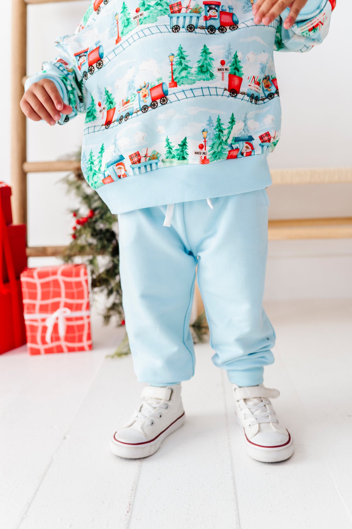 A toddler wearing a light blue sweatshirt and matching jogger pants with a festive Christmas train print, paired with white sneakers, standing in a brightly lit room with holiday decorations.