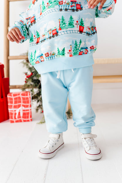 A toddler wearing a light blue sweatshirt and matching jogger pants with a festive Christmas train print, paired with white sneakers, standing in a brightly lit room with holiday decorations.