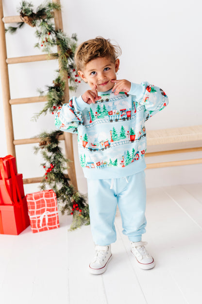 A young boy wearing a light blue Christmas-themed sweatshirt and matching sweatpants stands in a bright room with a wooden ladder, holiday garland, and wrapped gifts in the background.