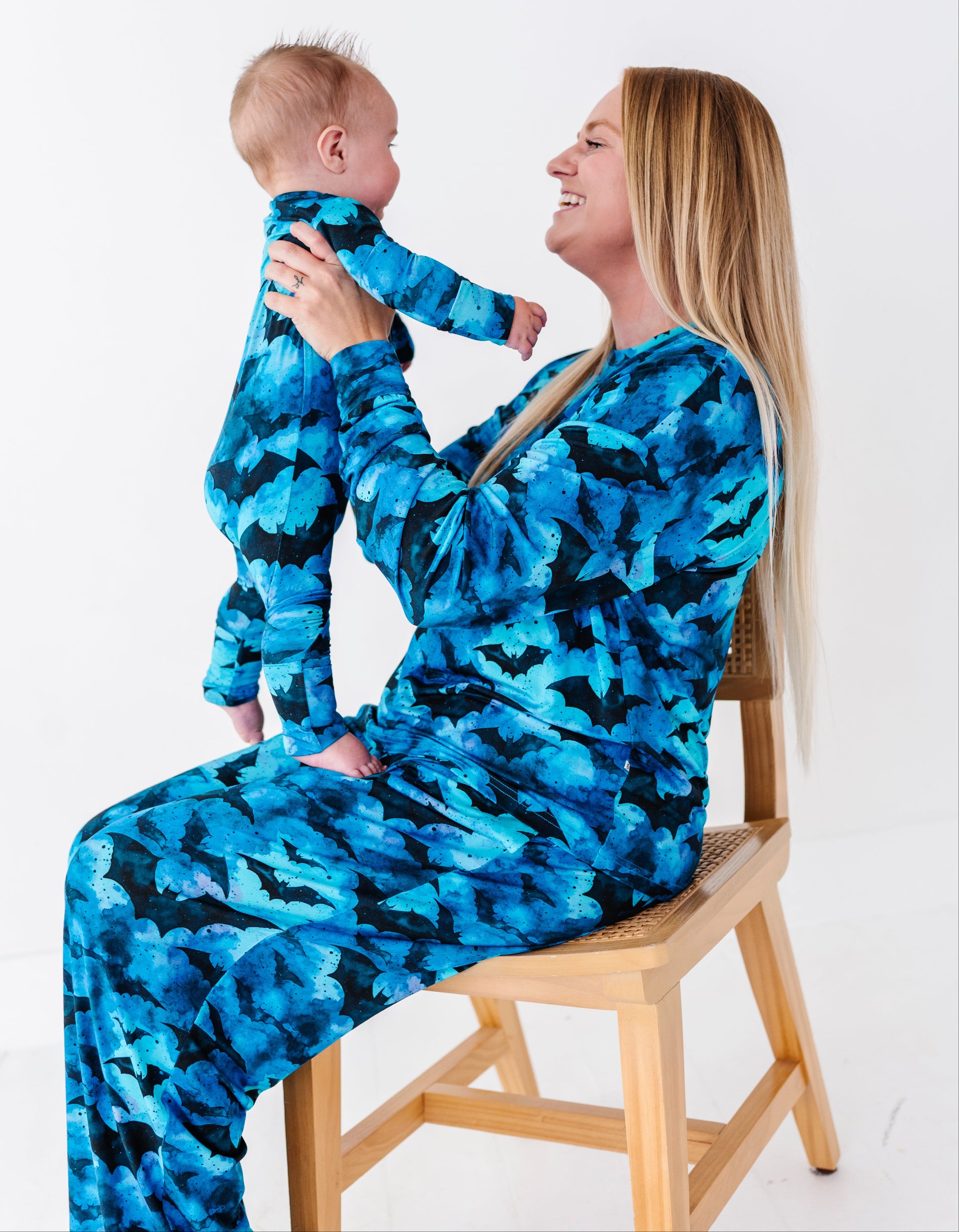 A woman and baby wearing matching blue and black patterned long-sleeve pajamas, sitting on a wooden chair against a white studio background.