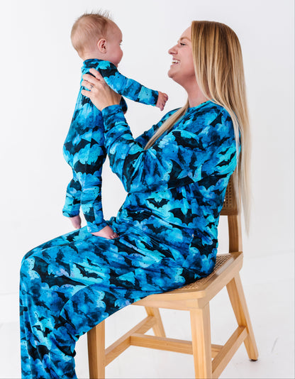 A woman and baby wearing matching blue and black patterned long-sleeve pajamas, sitting on a wooden chair against a white studio background.