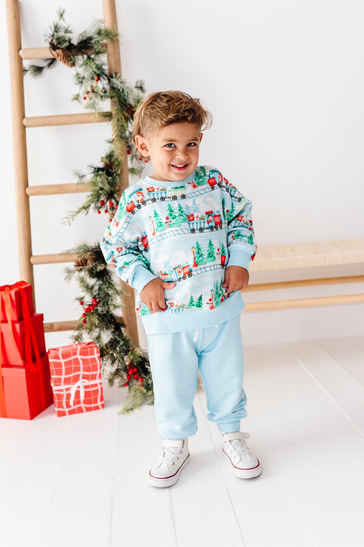 A young boy wearing a light blue Christmas-themed sweatshirt and matching sweatpants, standing in a bright room with a wooden ladder, holiday garland, and wrapped gifts.