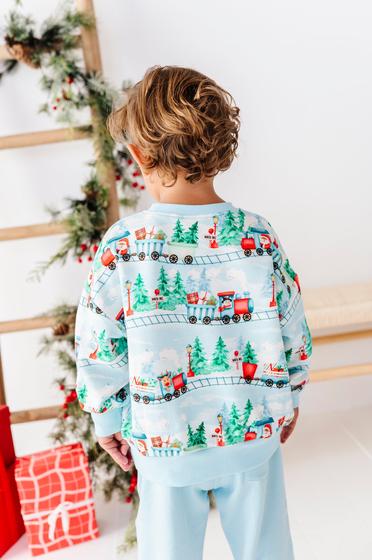 A child viewed from behind wearing a light blue Christmas-themed sweatshirt and matching pants, featuring a repeating pattern of red trains, green pine trees, and holiday gifts. The child is standing in a brightly lit room next to a decorated Christmas tree and wrapped presents.