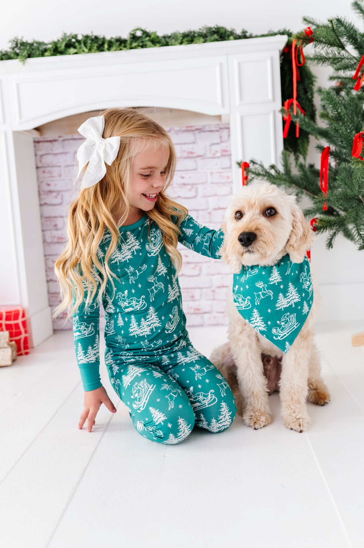 A blonde little girl wearing a bamboo two-piece pajama set. The pajamas feature vintage sleighs, Santas, reindeer, and pine trees. She's sitting with her dog in front of a fireplace and a ribbon-filled Christmas tree