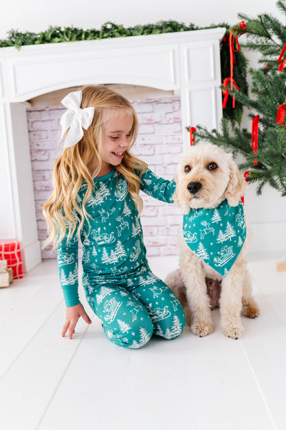 A blonde little girl wearing a bamboo two-piece pajama set. The pajamas feature vintage sleighs, Santas, reindeer, and pine trees. She's sitting with her dog in front of a fireplace and a ribbon-filled Christmas tree