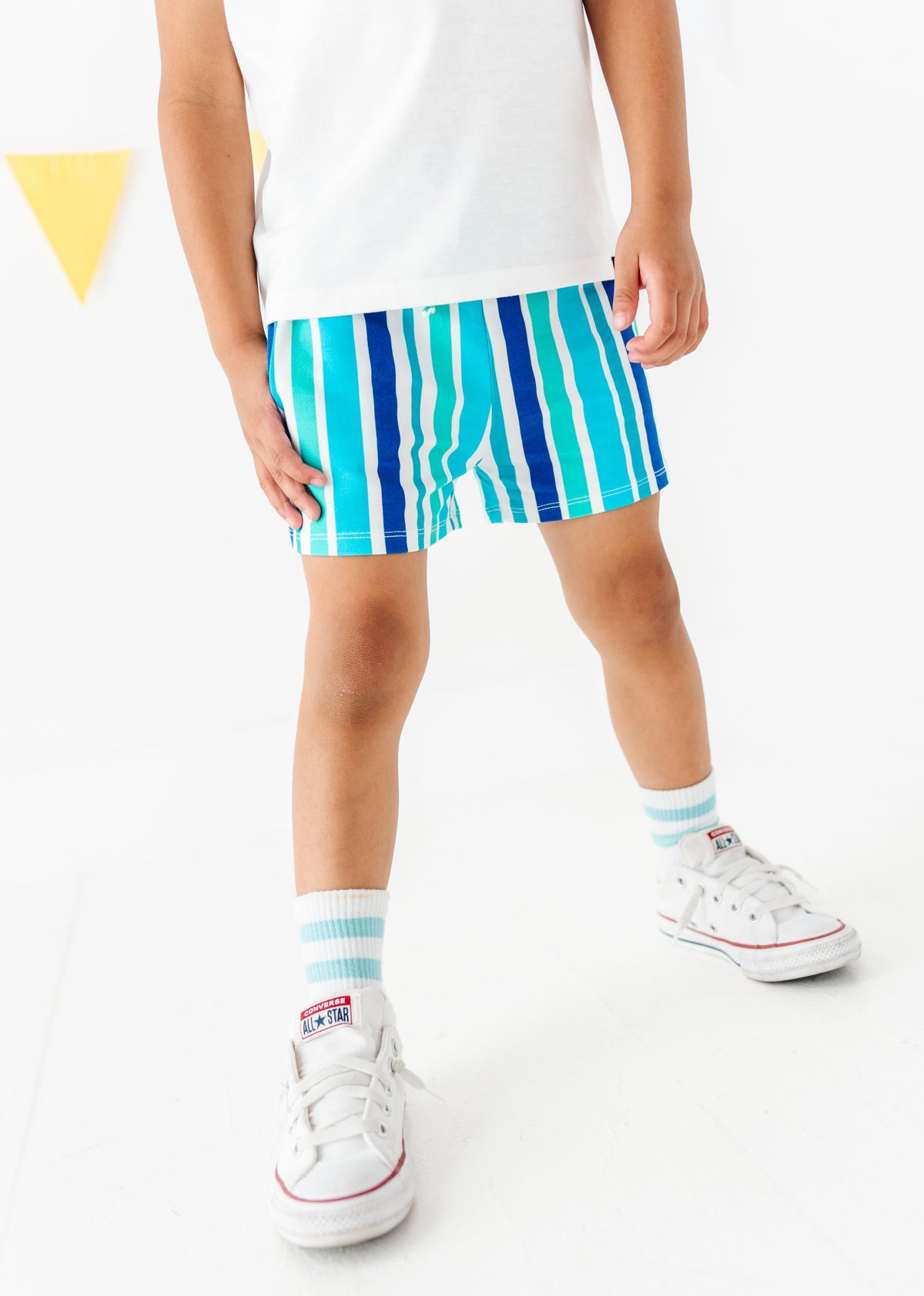 A child wearing striped boxer-brief style shorts in shades of blue, teal, and turquoise, paired with a white t-shirt, white socks, and white sneakers, shown from the waist down on a white studio background.