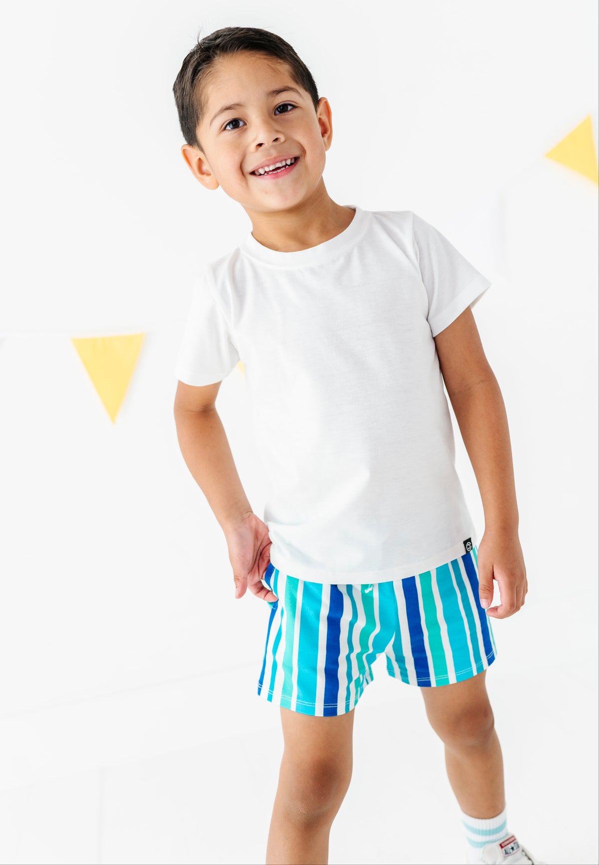 A young boy smiling and looking up, wearing a plain white short-sleeve t-shirt and blue and green vertically striped shorts against a white background with yellow triangle decorations.