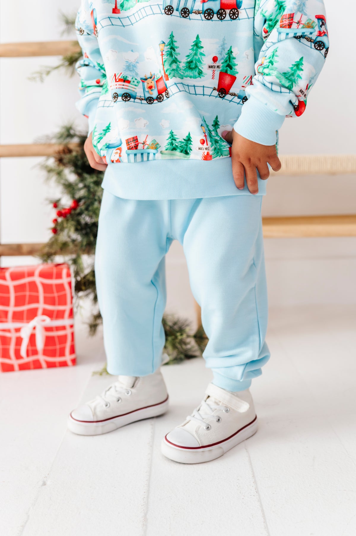 A child wearing a light blue sweatshirt and matching sweatpants with a Christmas train pattern, paired with white sneakers, standing in a festive indoor setting with a Christmas tree and gift box in the background.