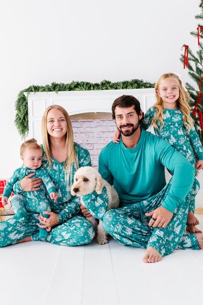 A family of four wearing matching teal and white holiday pajamas sits together on a white floor in front of a white fireplace mantel with green garland. A small white dog is held by the mother, and a Christmas tree is visible on the right.