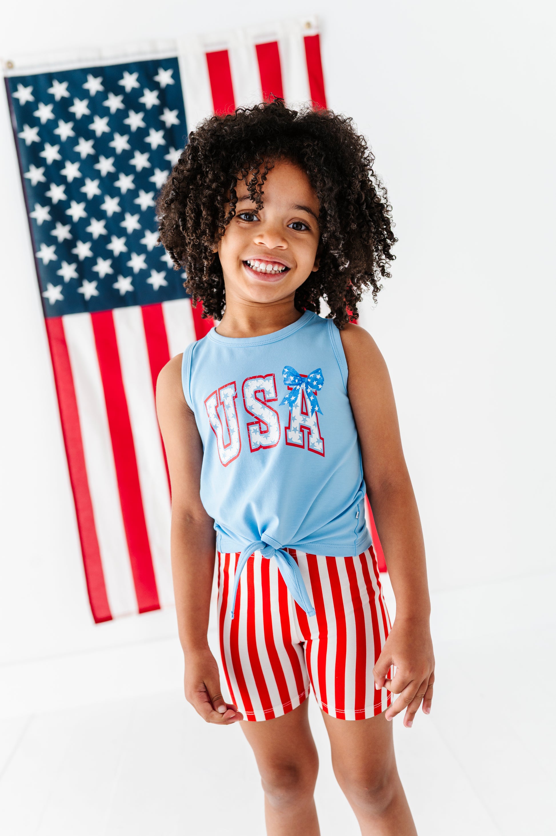 Child wearing a patriotic 'USA' bamboo tank top and striped bamboo biker shorts in front of an American flag.