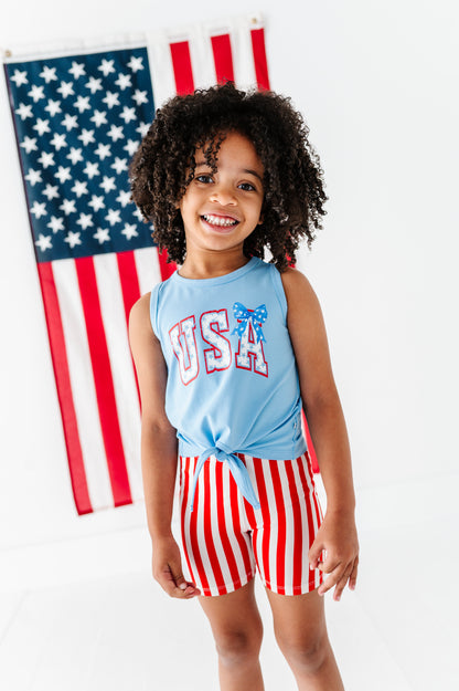 Child wearing a patriotic 'USA' bamboo tank top and striped bamboo biker shorts in front of an American flag.