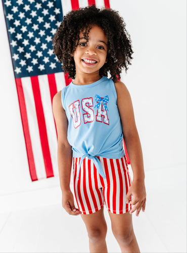 Child wearing a blue 'USA' top and red and white striped bamboo biker shorts in front of an American flag.