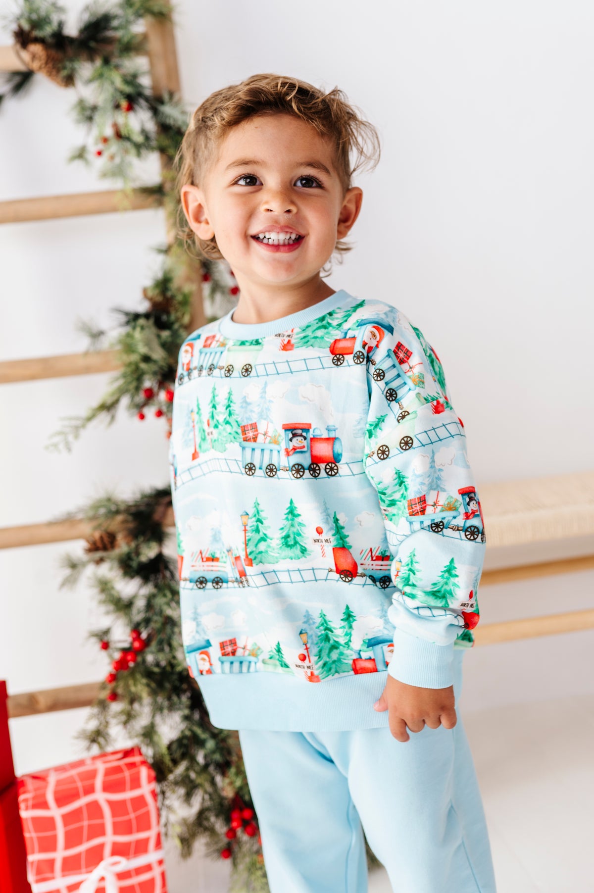 A smiling young boy wearing a light blue Christmas-themed crewneck sweatshirt and matching sweatpants, standing in front of a decorated Christmas tree and wooden ladder.