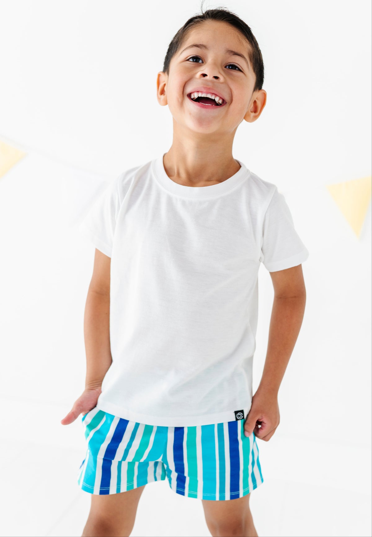 A young boy smiling and looking up, wearing a plain white short-sleeve t-shirt and blue and white vertically striped shorts against a bright white background.