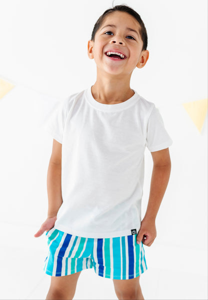 A young boy smiling and looking up, wearing a plain white short-sleeve t-shirt and blue and white vertically striped shorts against a bright white background.